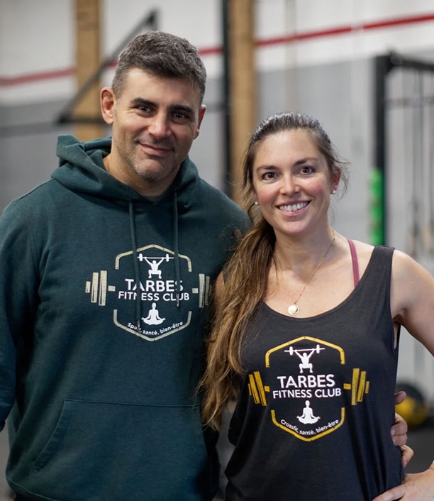 Couple souriant dans une salle de sport. L'homme et la femme portent des hauts Tarbes Fitness Club affichant 'Sport, santé, bien-être'.