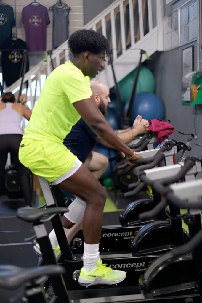 Un homme en tenue jaune fluo pédale intensément sur un vélo de spinning Concept 2 BikeErg dans une salle de sport, d'autres personnes s'entraînent.