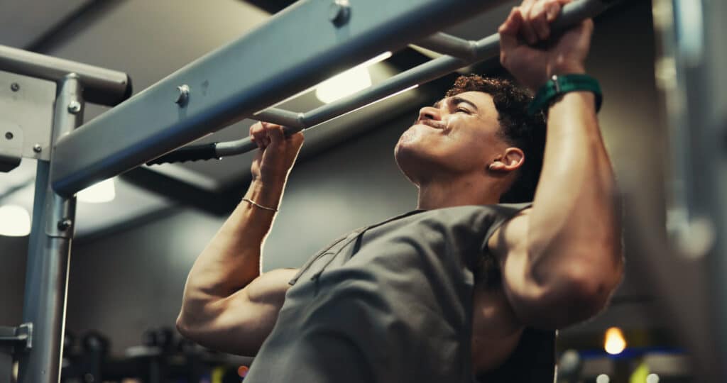 Homme athlétique en sueur faisant des tractions intensives sur une barre de musculation. Ses biceps sont contractés, visage concentré.
