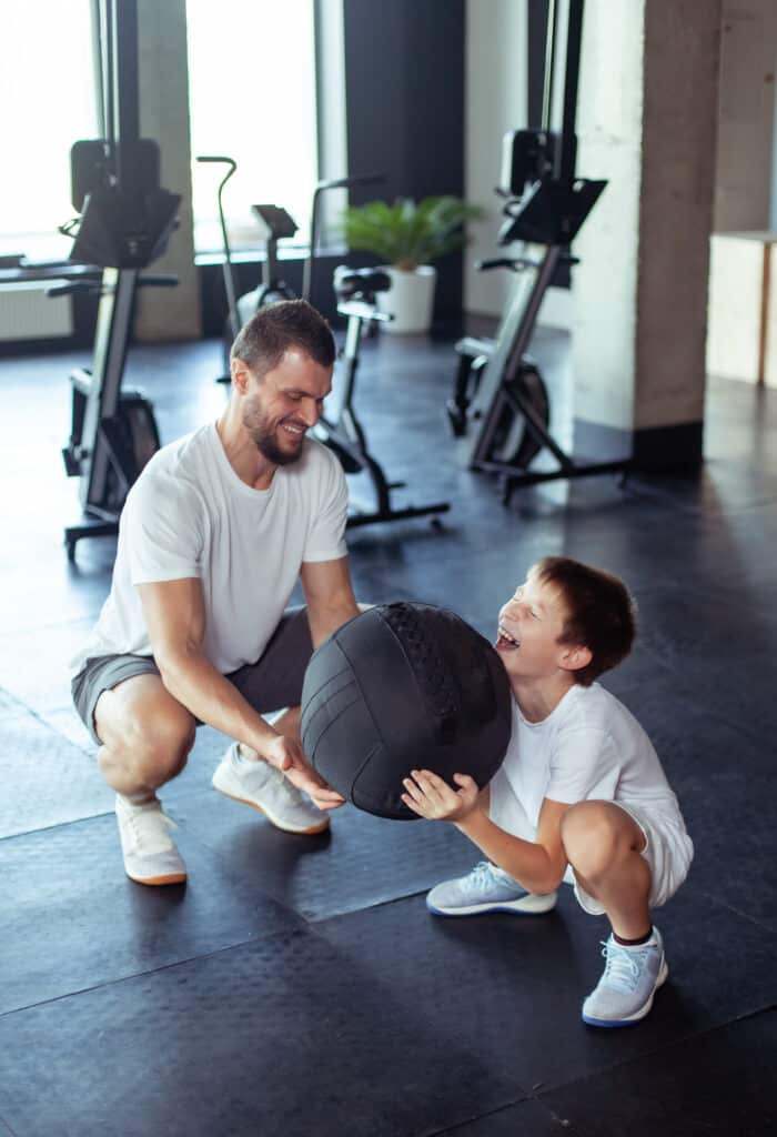 Père et fils joyeux s'entraînent au gym. L'homme sourit en aidant le garçon rieur à tenir un médecine-ball noir.