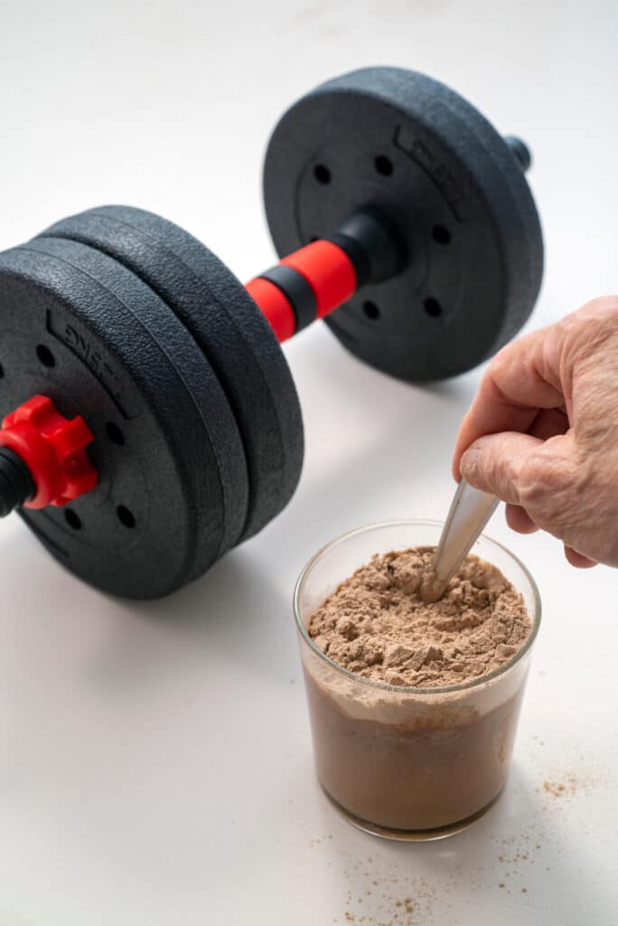 Main mélangeant de la poudre protéinée dans un verre, près d'un haltère noir et rouge de 2.5KG. Préparation après entraînement.
