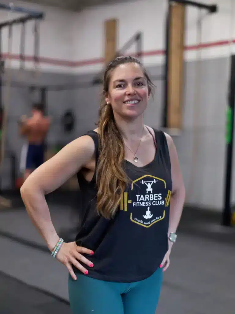 Femme souriante au Tarbes Fitness Club, débardeur noir avec logo or 'Crossfit, santé, bien-être' dans une salle de sport.