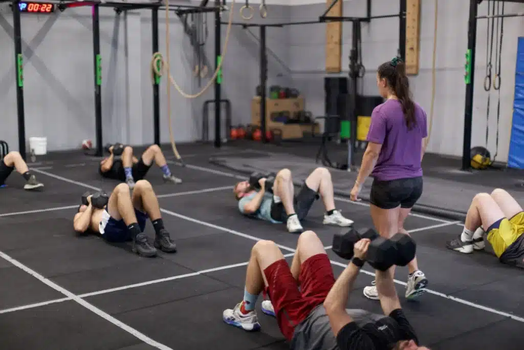 Plusieurs personnes s'entraînent au sol avec haltères dans un gymnase, supervisées par une coach. Un chrono affiche P 00:22.