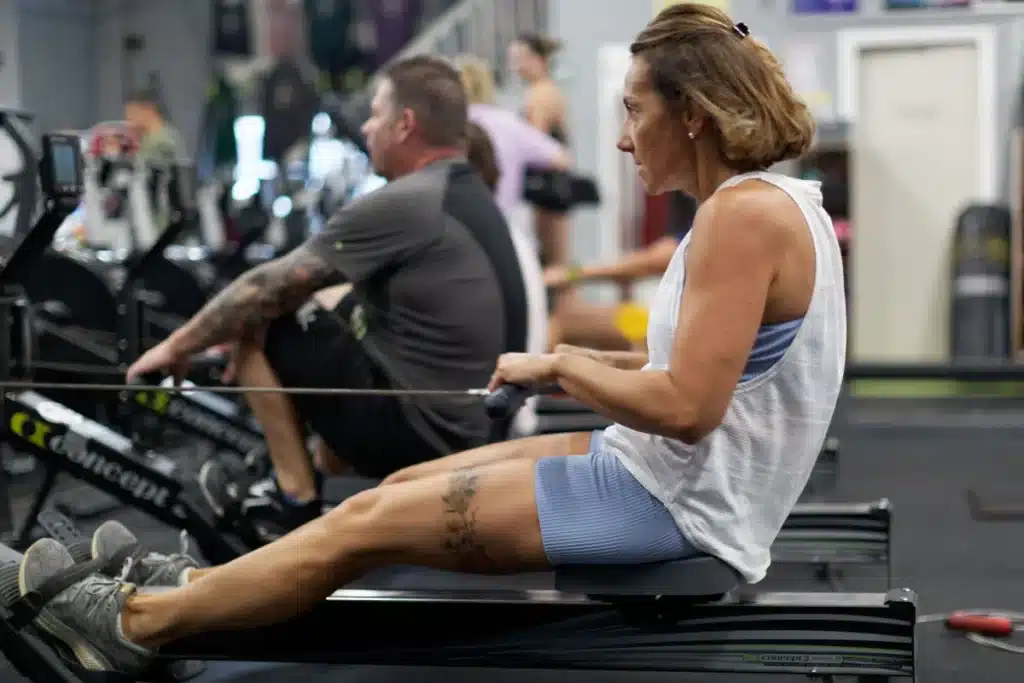 Femme concentrée rame sur un Concept2 en salle de sport, tatouage floral sur la cuisse. Un homme tatoué s'entraîne derrière.