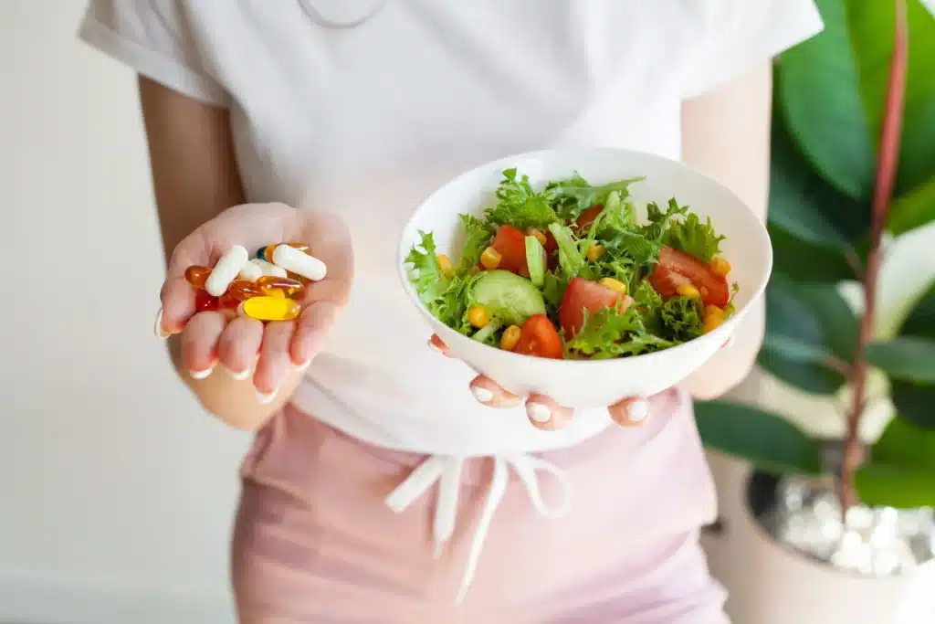 Personne tenant une poignée de gélules colorées et un bol de salade fraîche. Représente le choix entre suppléments et alimentation saine.