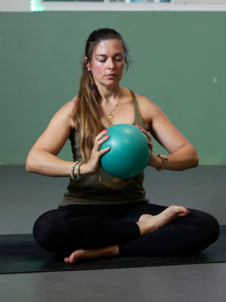 Pilates : Méditation et exercice avec ballon Femme en tenue de sport, assise en tailleur sur un tapis, les yeux fermés, tenant un petit ballon de Pilates bleu-vert.